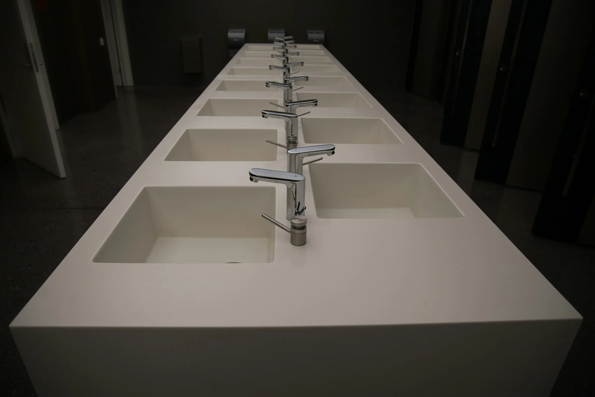 Long white washroom counter with a row of built-in square basins and chrome taps